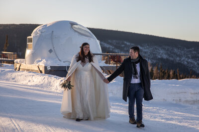 Bride and groom walk holding hands in the snow, an igloo airbnb sits in the background of this couples Northern Lights elopement