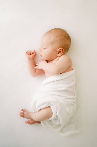 Newborn baby boy lays on his side wrapped in white swaddle during studio photography session in Raleigh NC