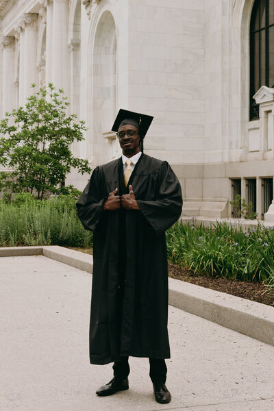 Photo of a woman sitting on the ledge of a building with her feet dangling with more college buildings in the background behind her. She is wearing her graduation cap and sash