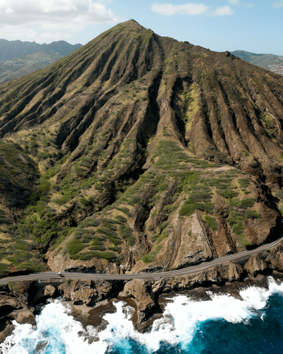 a large mountainscape in hawaii rises up above a winding coastal road abutting the sea