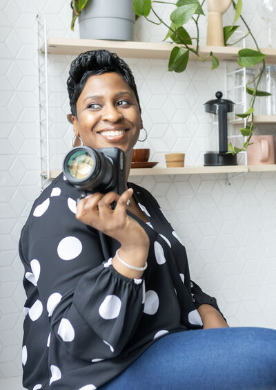 Metro Detroit Senior Photographer  with short pixie haircut in black polka dot blouse and blue jean jeggings posing casually holding camera