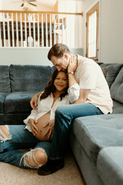 Couple sitting on Lake Michigan beach. The woman is holding her pregnant belly and man has his arms wrapped around her and is kissing her on the forehead.