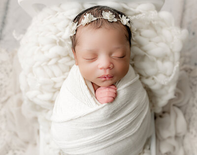 Newborn Photographer, a baby girl wheres a flower head band and is wrapped in white linens