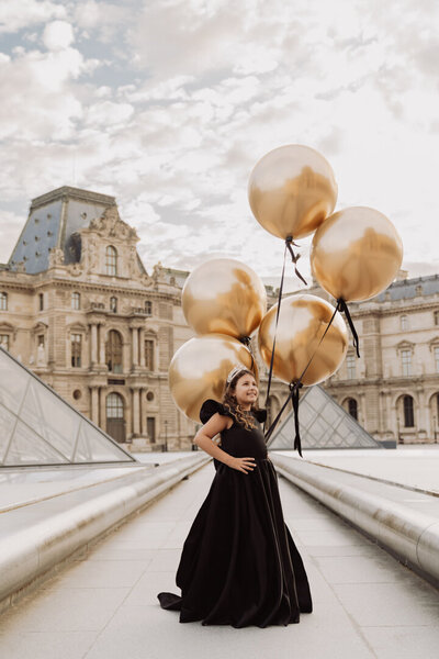 Little girl in black princess dress holding golden balloons during Louvre Paris photoshoot