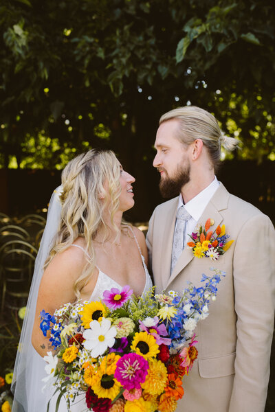 Bride and groom at Ojai Rancho Inn/ The bride is holding a colorful wildflower bouquet with sunflowers and the groom has a floral pocket square and a beige groom's suit with gilded shoes.
