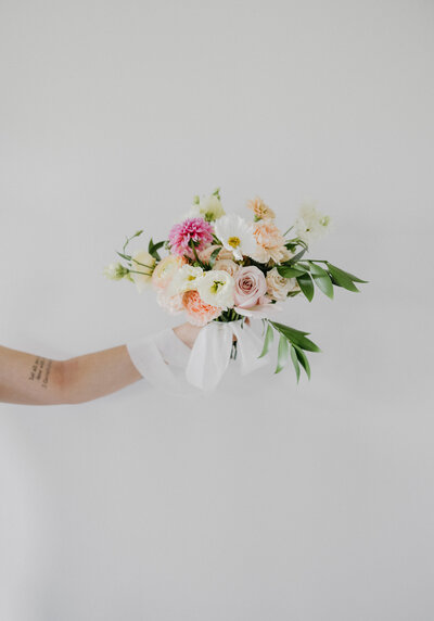 Hand holding a pastel garden-style bridesmaid bouquet featuring blush roses, soft peach ranunculus, white lisianthus, pink dahlias, and delicate greenery with flowing white ribbon, photographed against a clean light gray backdrop for a modern, airy wedding aesthetic.