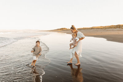A mother holds her baby while her son plays in the water at the beach during a family photoshoot in canterbury