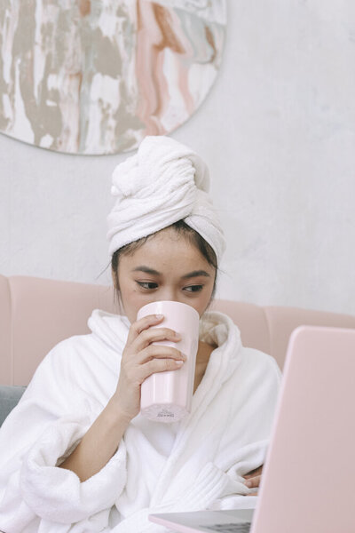 A young Asian woman sits on a light pink couch, wearing a robe and a towel around her head. She is taking a drink from a pink tumbler, looking down toward a laptop in front of her.