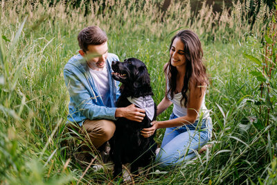 Couple snuggles their black  retriever amongst a field of tall grasses