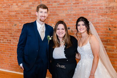 A man in a blue suit and two women, one in a white wedding dress and the other in a black outfit, smiling together in front of a brick wall at a Quad Cities wedding.