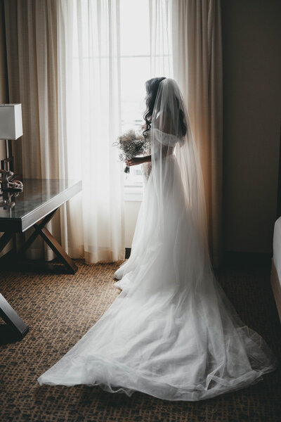 bride standing next to a window holding wedding bouquet