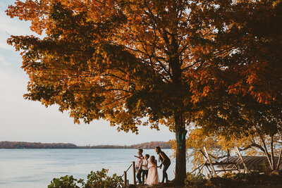 family is standing on edge of lake, pointing out to the water, while standing under a big oak tree in the fall with beautiful red leaves