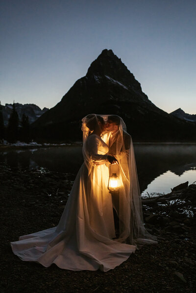 A bride and groom share an intimate kiss under a bridal veil illuminated by a lantern beside a mountain lake at dusk in Glacier National Park, captured by Sydney Breann Photography.