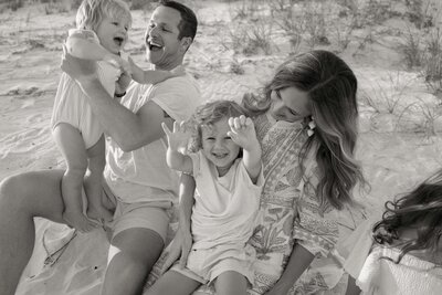 Black and white photo of mom and dad laughing with their two small children sitting on the beach