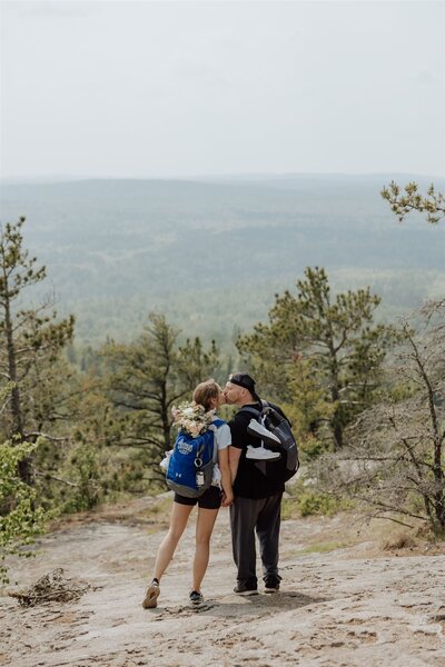couple in hiking attire wearing backpacks kissing on a mountaintop