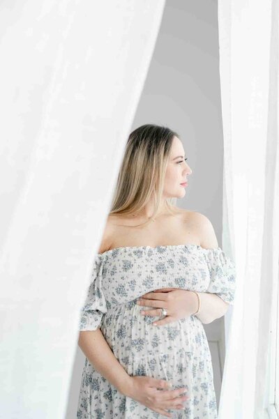 Pregnant woman in white floral dress stands in front of a window.