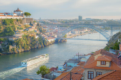 Scenic view of a river in Porto, Portugal, with a cruise boat sailing under the iconic Dom Luís I Bridge. The foreground shows traditional terracotta-roofed buildings, while the hillside and cityscape extend in the background.