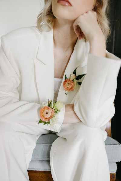 A modern, fashion-forward bride wearing an elegant white tailored suit sits thoughtfully with her arm resting on her knee, showcasing two delicate floral corsages made of coral ranunculus, white blooms, and greenery pinned to her lapel and sleeve—minimalist bridal styling featuring contemporary wedding florals in soft peach and cream tones.