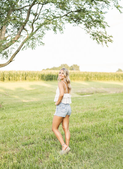 high school senior female, with long blonde hair, standing in the grass with a tree and cornfield behind her, looking at the camera smiling, wearing blue jean shorts, sandals, and a white spaghetti strapped shirt