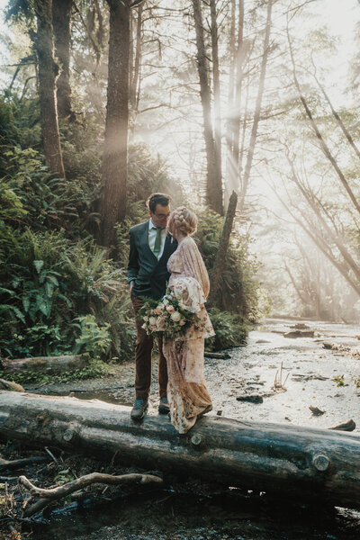 Bride wearing pink wedding dress and flowers in hair faces groom wearing green suit jacket looking into each other's eyes in Fern Canyon surrounded by light rays and trees taken by California elopement photographer Kasey Mantiply