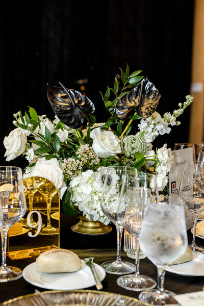 A glass, clear compote vase adorned with blush and white florals  in the middle of a table featuring a clear charger plate with gold rim and a white dinner plate with gold rim  with gold silver ware with clear handle