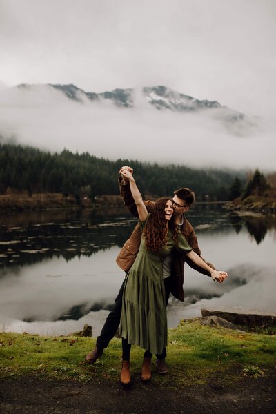 man and woman stand holding hands arms outstretched with lake and mountain behind them
