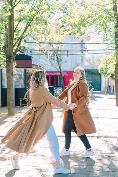 Lesbian couple dancing and holding hands while smiling at each other on rue de l’Abreuvoir in Montmartre, both wearing long beige jackets during a romantic Paris photoshoot.