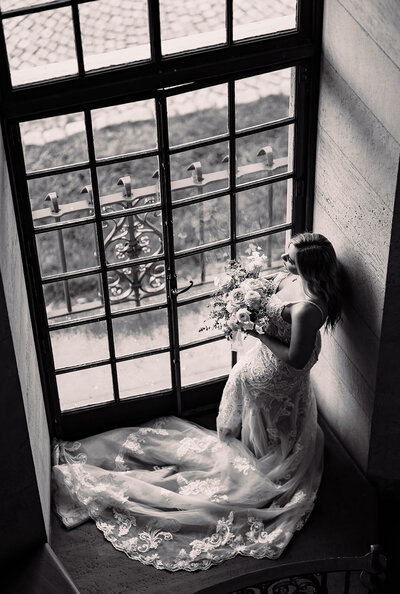 Bride holding her bouquet staring out a window with the photo taken from above her. Her train is wrapped around her feet.