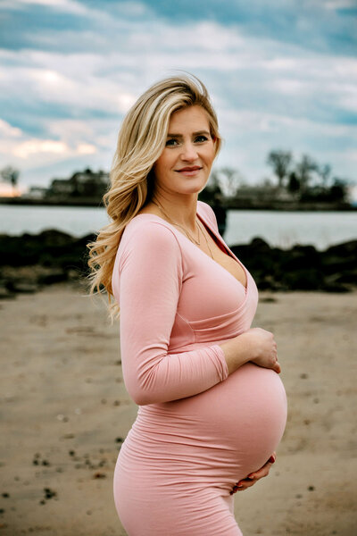 Mom-to-be in pink gown standing near a lake in Stamford, CT for her maternity session.