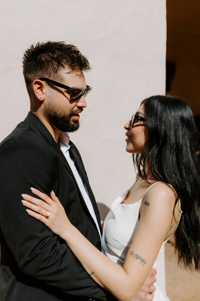 A stylish couple stands close to each other in warm sunlight, both wearing sunglasses and sharing a quiet moment.