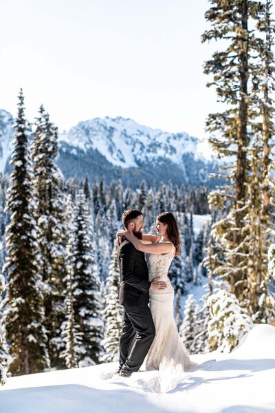 bride and groom are standing on a rock with a vast sky background and mountains behind them. They are kissing and her white dress is blowing in the wind. She is holding a bouqet with yellow sunflowers