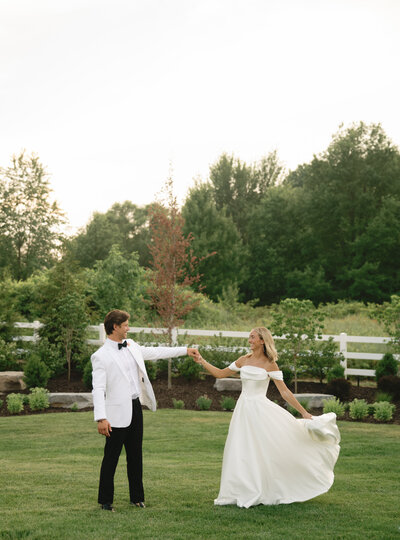 A groom in a white jacket and his bride dance out side  by a white fence on their wedding day at Venue Bella Giornata in Michigan.