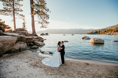 Bride and groom embracing on the sandy shoreline at Sand Harbor in Lake Tahoe, with calm blue water, large granite rocks, and snow-capped mountains in the distance at sunset.