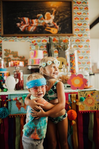 Two children dressed like hippies standing in a room of flower crafts hugging each other