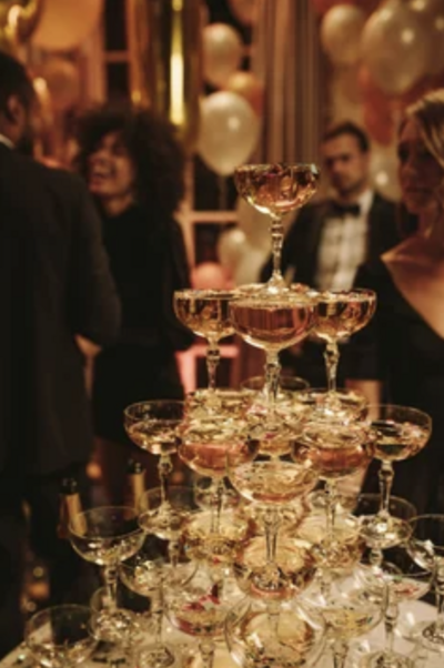 A luxurious party setup with a champagne tower surrounded by guests. The scene includes festive balloons and an elegant atmosphere.