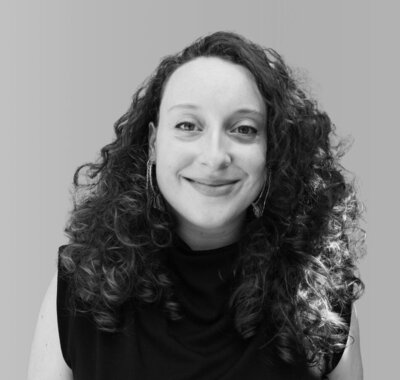 Black and white photo of woman with curly brown hair smiling at the camera wearing a black turtleneck and detailed earrings.