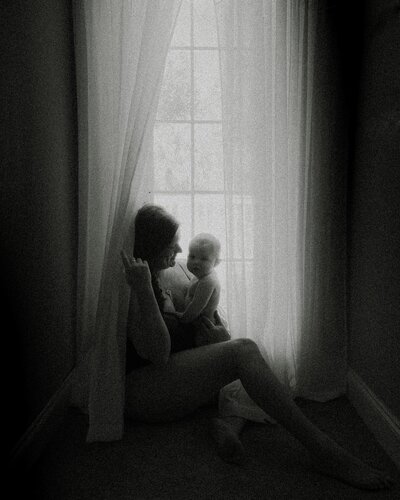 A mother and baby sit by a window. The mother is looking at the baby, the baby is looking at the camera while in their mother's arms. The mother is holding the window curtain