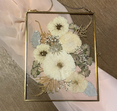 Elegant pressed floral arrangement featuring white roses and baby's breath in a gold frame, displayed on a soft pink fabric background. Perfect for preserving special memories