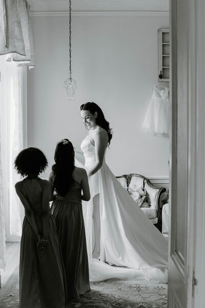 Rhode Island Wedding Photographers | A bride in an elegant gown stands in a softly lit room, with two bridesmaids admiring her from behind. 