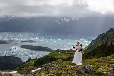 Bride and groom embrace on a dramatic mountaintop overlooking an Alaskan fjord filled with floating icebergs. The couple stands on a lush green ridge as sunlight breaks through the clouds, capturing the wild beauty of an adventurous Alaska elopement.