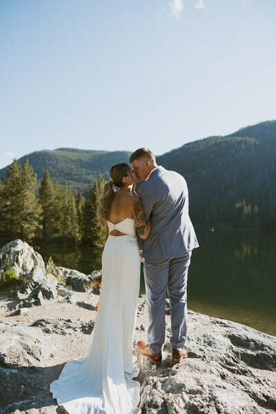 A couple who eloped at Officers Gulch Pond in Colorado, facing away from the camera, sharing a kiss with a stunning lake in front of them and mountains in the background, capturing the beauty of their intimate moment in nature.