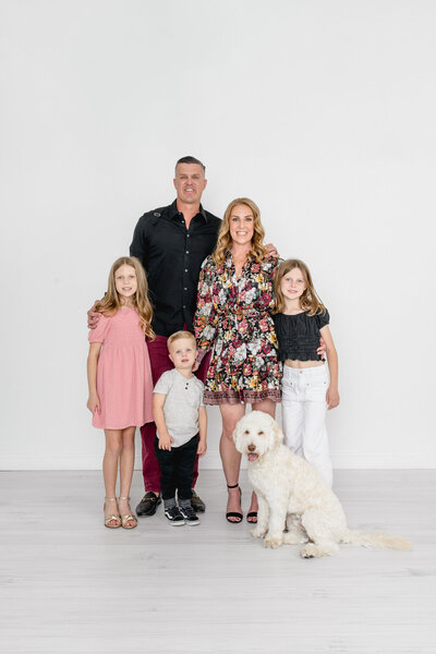 A family of five dressed in neutrals smiling at the camera with their white dog in a white studio