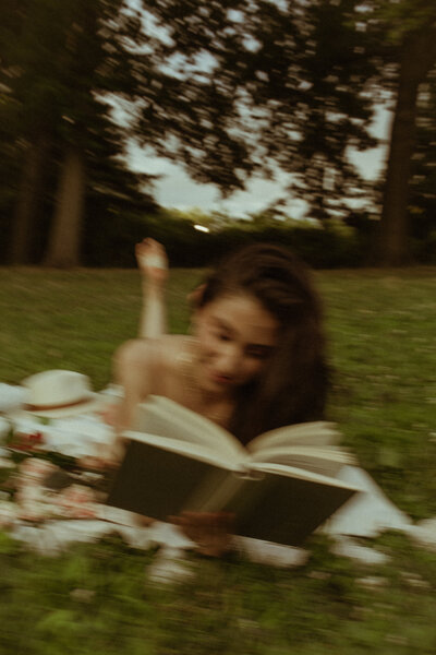 Moving shot of a woman lying on her stomach on a picnic blanket in the park  with her feet up and reading a book