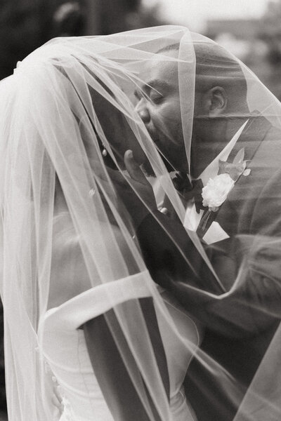 A newlywed couple kisses underneath the bride's veil in a nostalgic black and white photo.