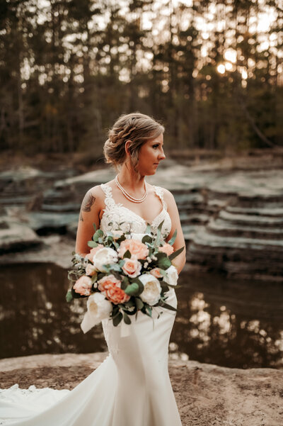 bride in a lace and satin wedding dress holding a white and cream wedding bouquet in little rock arkansas with a lake and unique rocks behind her in the woods