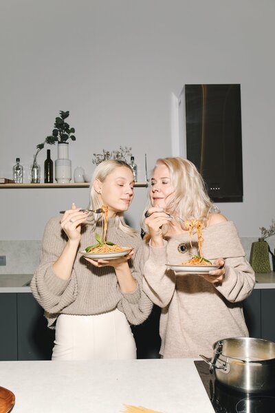 Two blonde women eating spaghetti. mother and daughter enjoying a home cooked meal in their kitchen. Both women are wearing tan oversized knit sweaters.