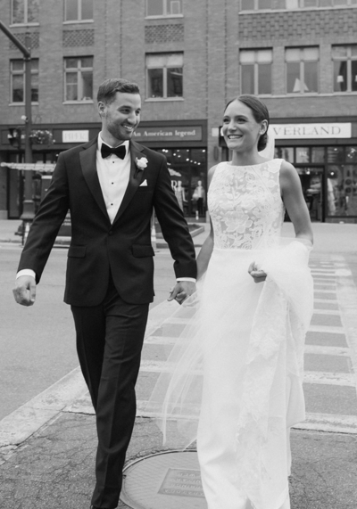 Black and white image of bride and groom holding hands walking across a city street crosswalk