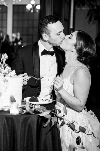 In a candlelit room, a couple in formal attire shares a kiss beside a beautifully adorned cake on a small table. The white cake, decorated with delicate florals, complements the romantic ambiance of their memorable Lilah Philadelphia wedding.