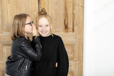 two tween sister, with one whispering into the other girls ear while she giggles looking at the camera