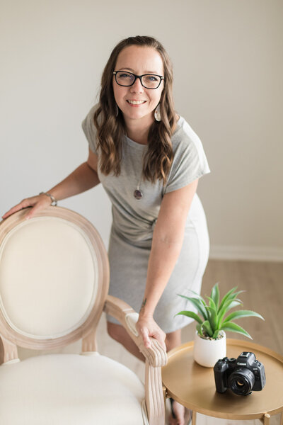 Dark brunette woman standing next to a chair wearing a gray dress, black glasses, and a table beside the chair with a green plant and black camera